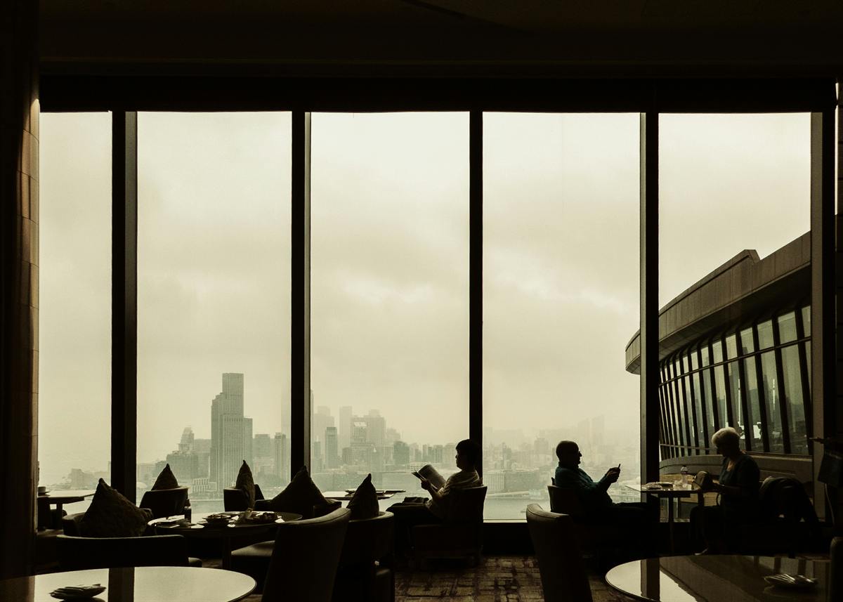 People reading and relaxing in a dim lounge, silhouetted against floor-to-ceiling windows and a misty city skyline.