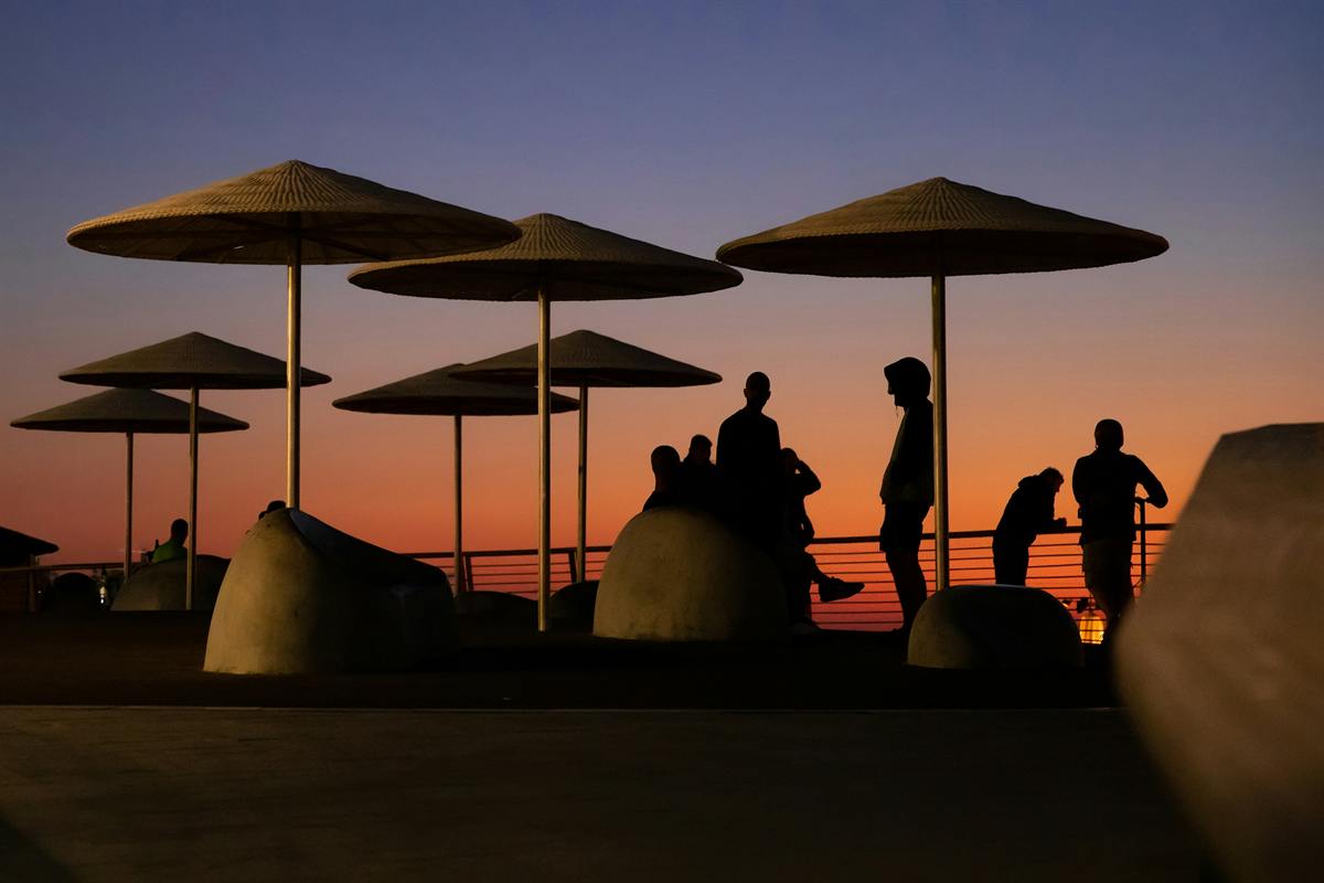 People silhouetted beneath large woven umbrellas on a seaside terrace at sunset.