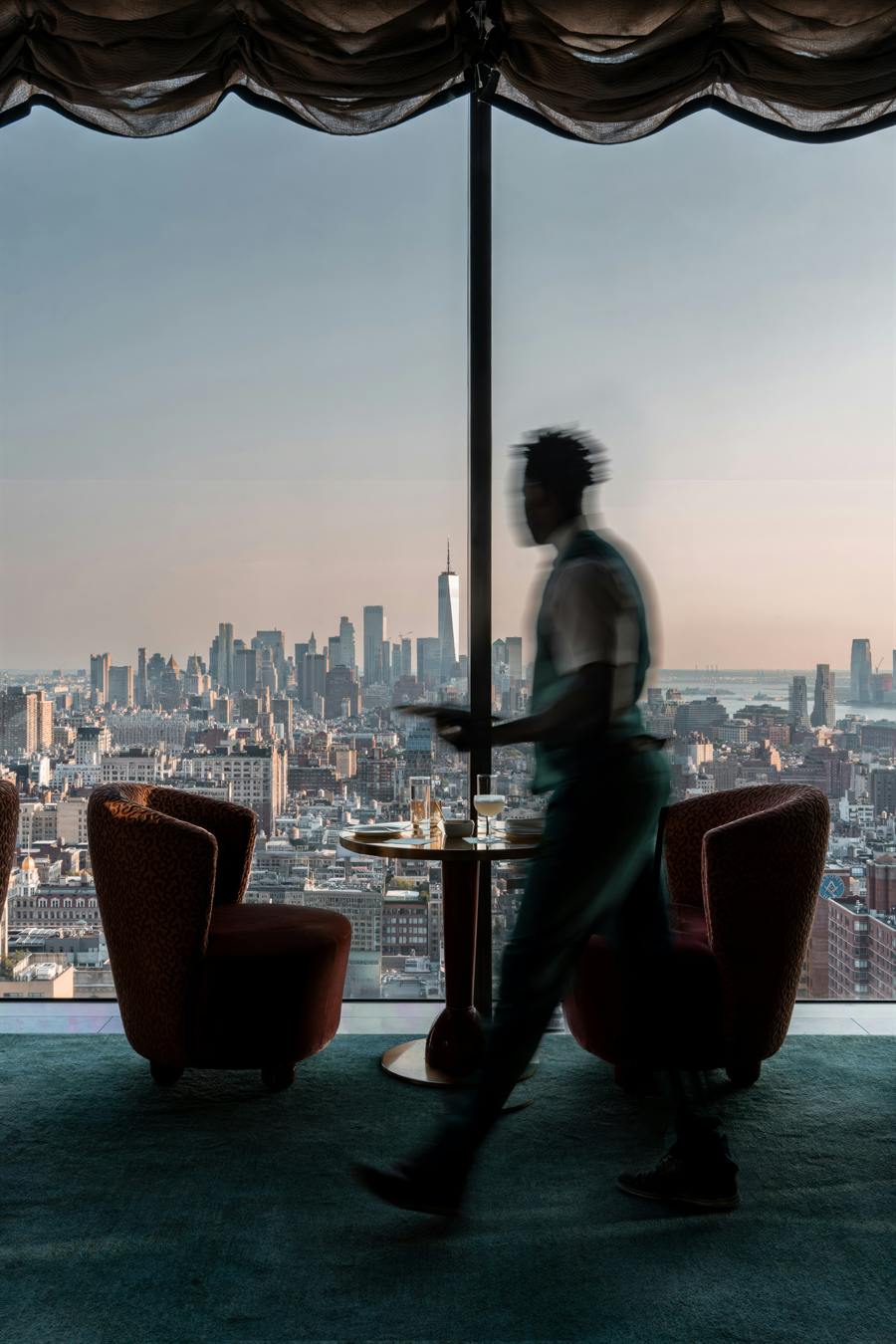 Two velvet lounge chairs and a small table beside a tall window overlooking a city skyline, as a blurred figure walks past.